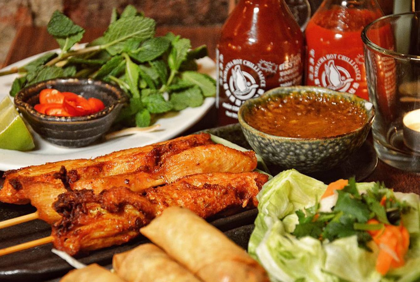 Char-grilled marinated chicken satay skewers and crispy spring rolls on a dark plate, with fresh mint, lime, lettuce, sliced chilli, peanut dipping sauce and bottles of chilli sauce behind.
