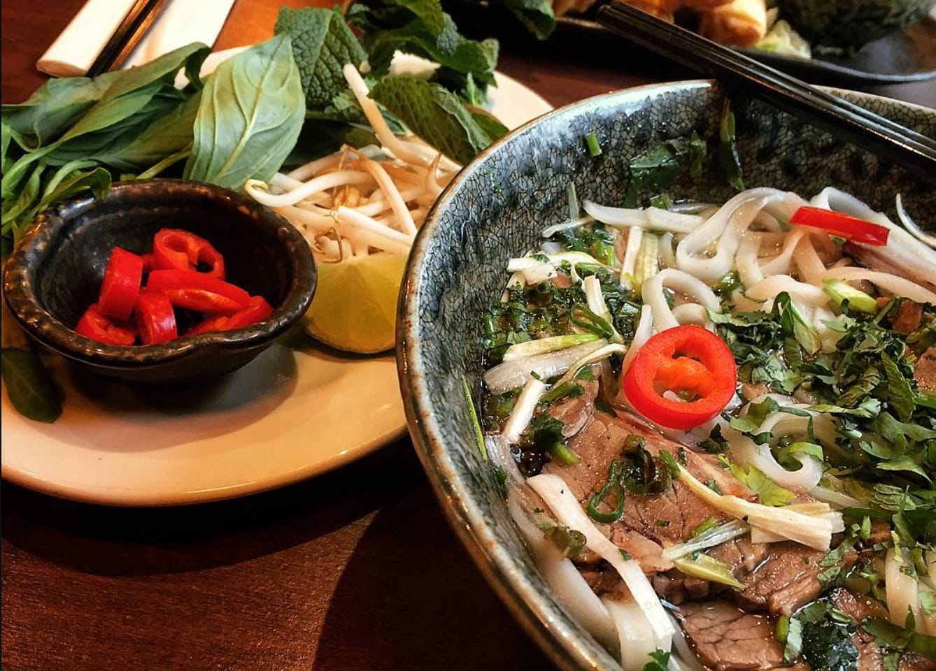 A bowl of phở — sliced beef and flat rice noodles in clear aromatic broth topped with coriander and spring onion, with a side plate of Thai basil, mint, beansprouts, lime and fresh red chilli.