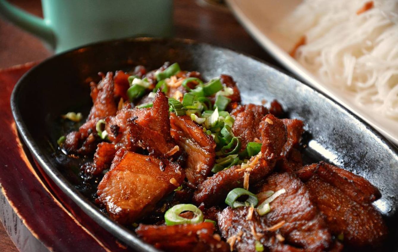 Caramelised slices of pork belly on a hot cast-iron plate, scattered with spring onion and fried garlic, served alongside a plate of rice vermicelli noodles.